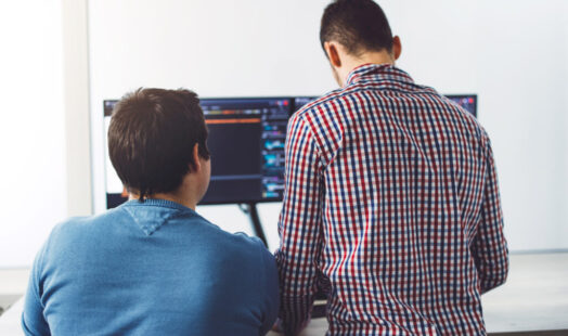 Back view of two young IT guys in front of computer screens, writing a new code Back view of two young IT guys in front of computer screens, writing a new code